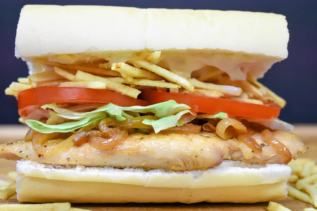 A thin chicken breast on Cuban bread topped with shredded lettuce, sliced tomatoes and potato sticks, on a wood cutting board.