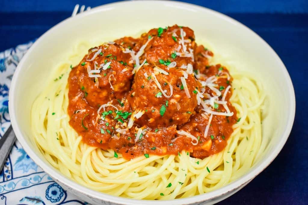 Italian sausage meatballs in a red sauce, served on a bed of spaghetti in a white bowl on a blue place mat and garnished with chopped parsley and grated Parmesan cheese.
