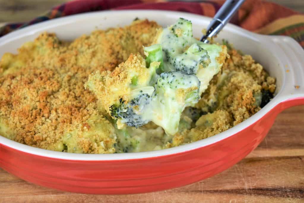 A large spoon scooping broccoli covered with creamy cheese and breadcrumbs that's served in a red and white casserole dish.