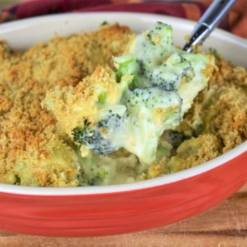 A large spoon scooping broccoli covered with creamy cheese and breadcrumbs that's served in a red and white casserole dish.