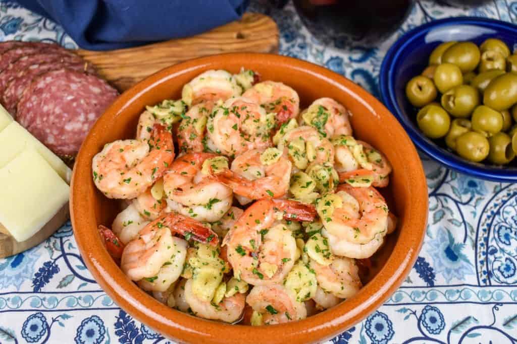 Camarones al ajillo served in an orange clay bowl with salami, cheese and olives in the background.