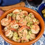 Camarones al ajillo served in an orange clay bowl with salami, cheese and olives in the background.