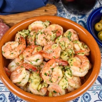 Camarones al ajillo served in an orange clay bowl with salami, cheese and olives in the background.