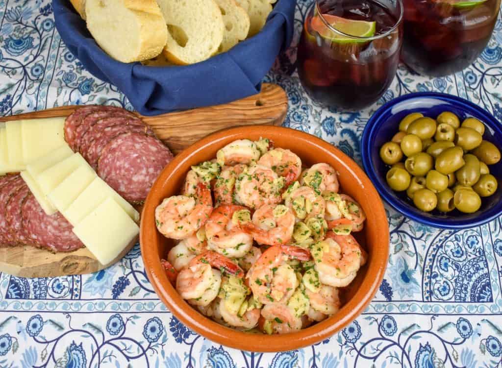 Camarones al ajillo served in an orange clay bowl with salami, cheese, olives, sliced bread and sangria served in the background.