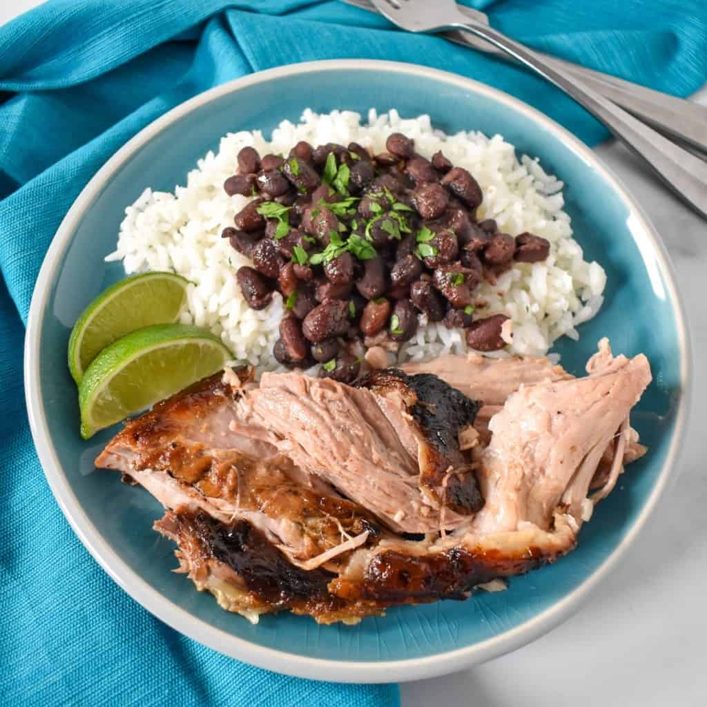 Pieces of the Cuban roast pork served with white rice and black beans on an aqua plate with an aqua linen and garnished with two lime wedges.
