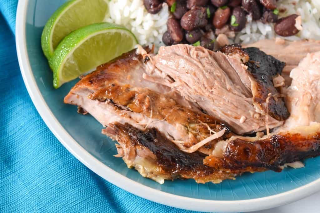 A close up image of pieces of roast pork on an aqua plate and linen, served with white rice and black beans and two lime wedges.