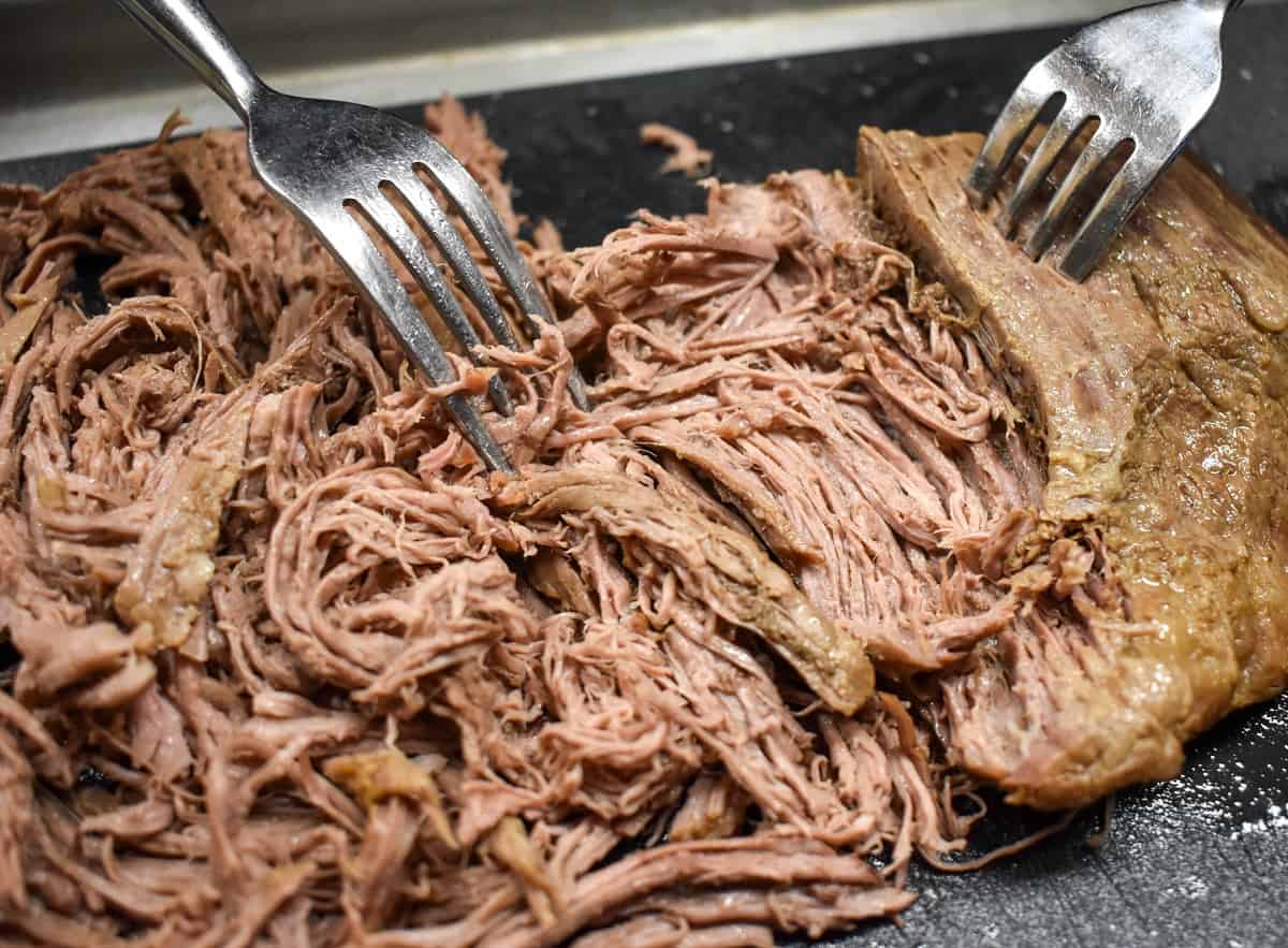 Cooked flank steak being shredded by two forks on a black cutting board.