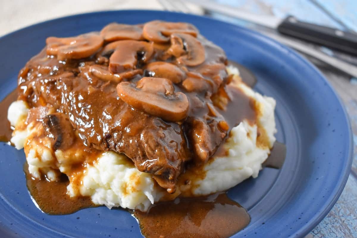 A thick sirloin steak covered in a dark brown mushroom gravy on a bed of mashed potatoes served on a blue plate.