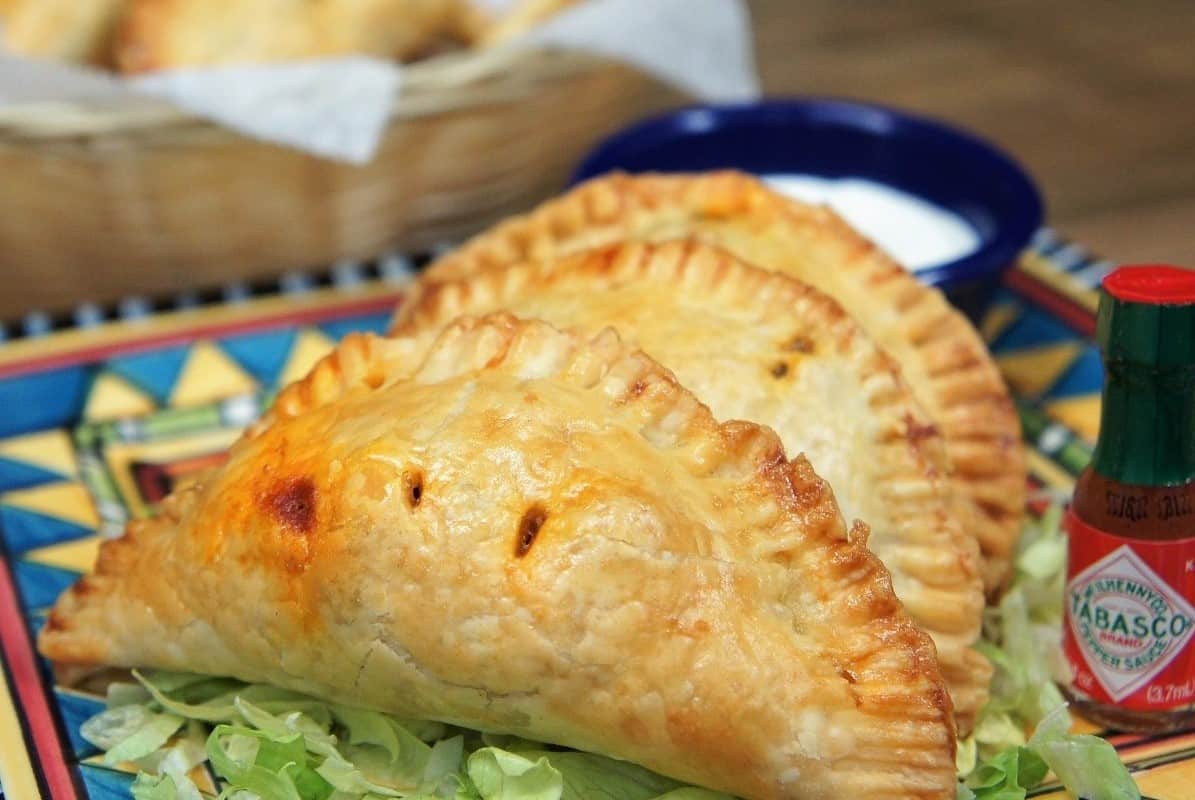 Taco Hand Pies three small hand pies on a colorful plate on a bed of shredded lettuce with hot sauce and sour cream on the side