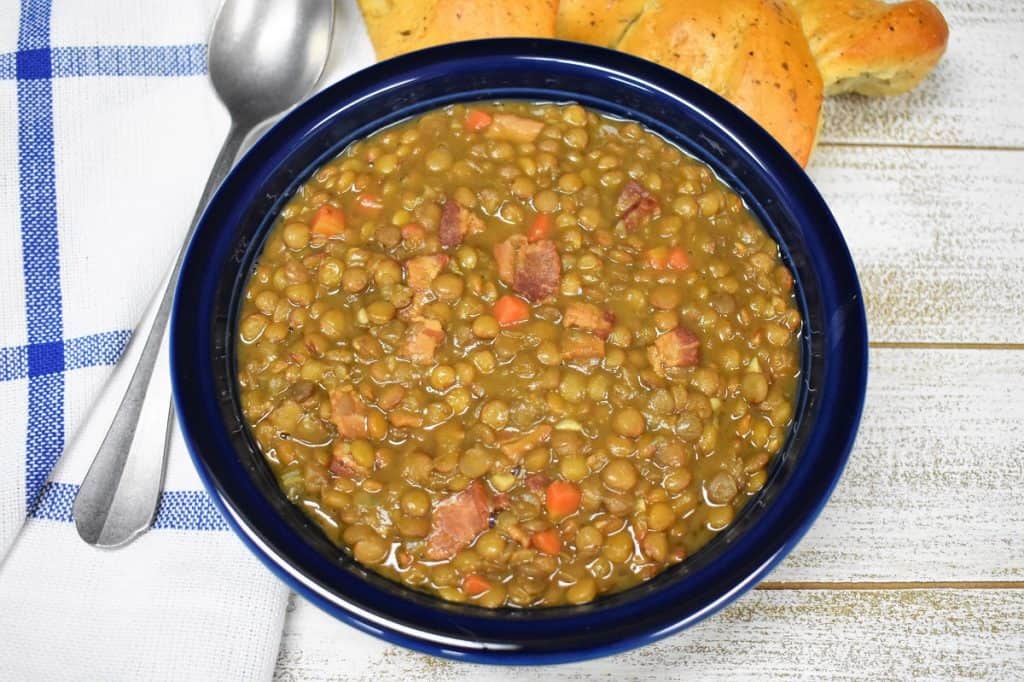 Bacon and lentil soup served in a blue plate with a piece of bread in the background and a spoon to the left.