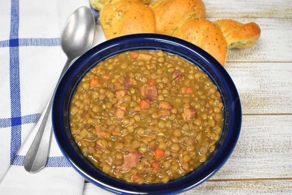 Bacon and lentil soup served in a blue bowl with a piece of bread in the background and a spoon to the left.