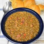 Bacon and lentil soup served in a blue bowl with a piece of bread in the background and a spoon to the left.