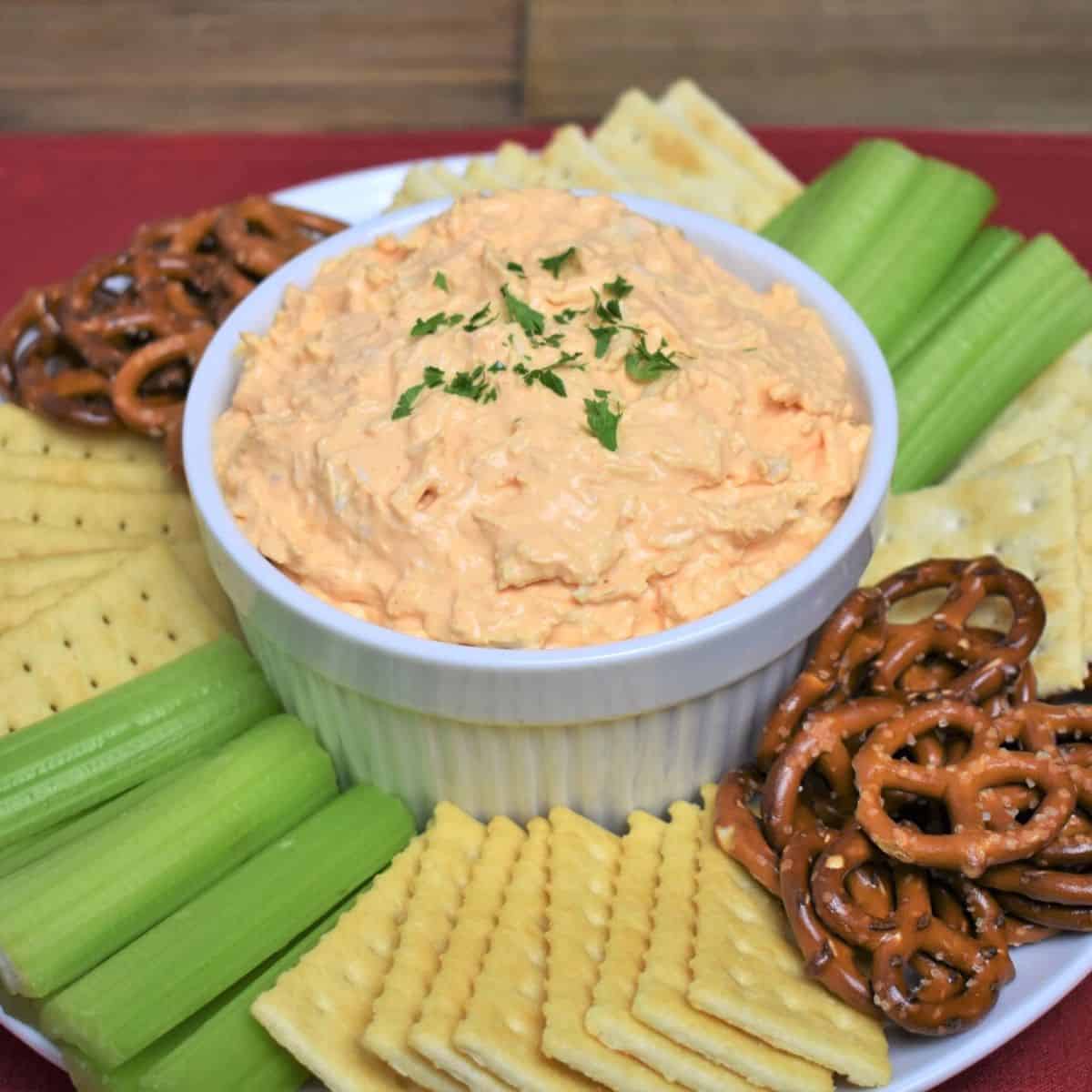 Cold Buffalo Chicken Dip served in a white bowl with assorted crackers, celery sticks and pretzels arranged on the plate.