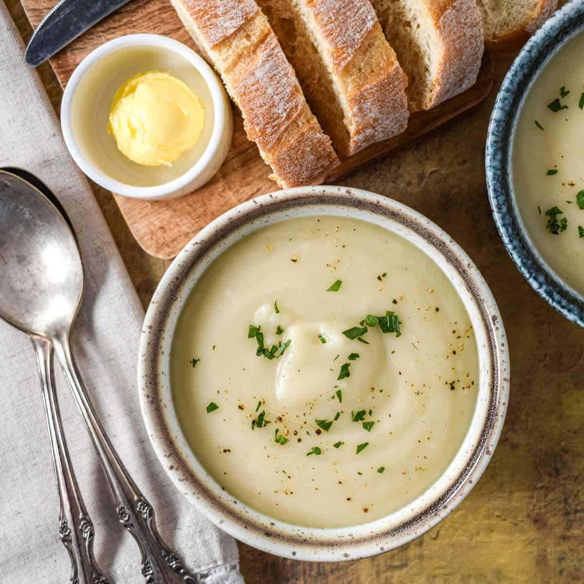 Close-up of a bowl of creamy soup topped with chopped parsley, with bread and butter in the background.
