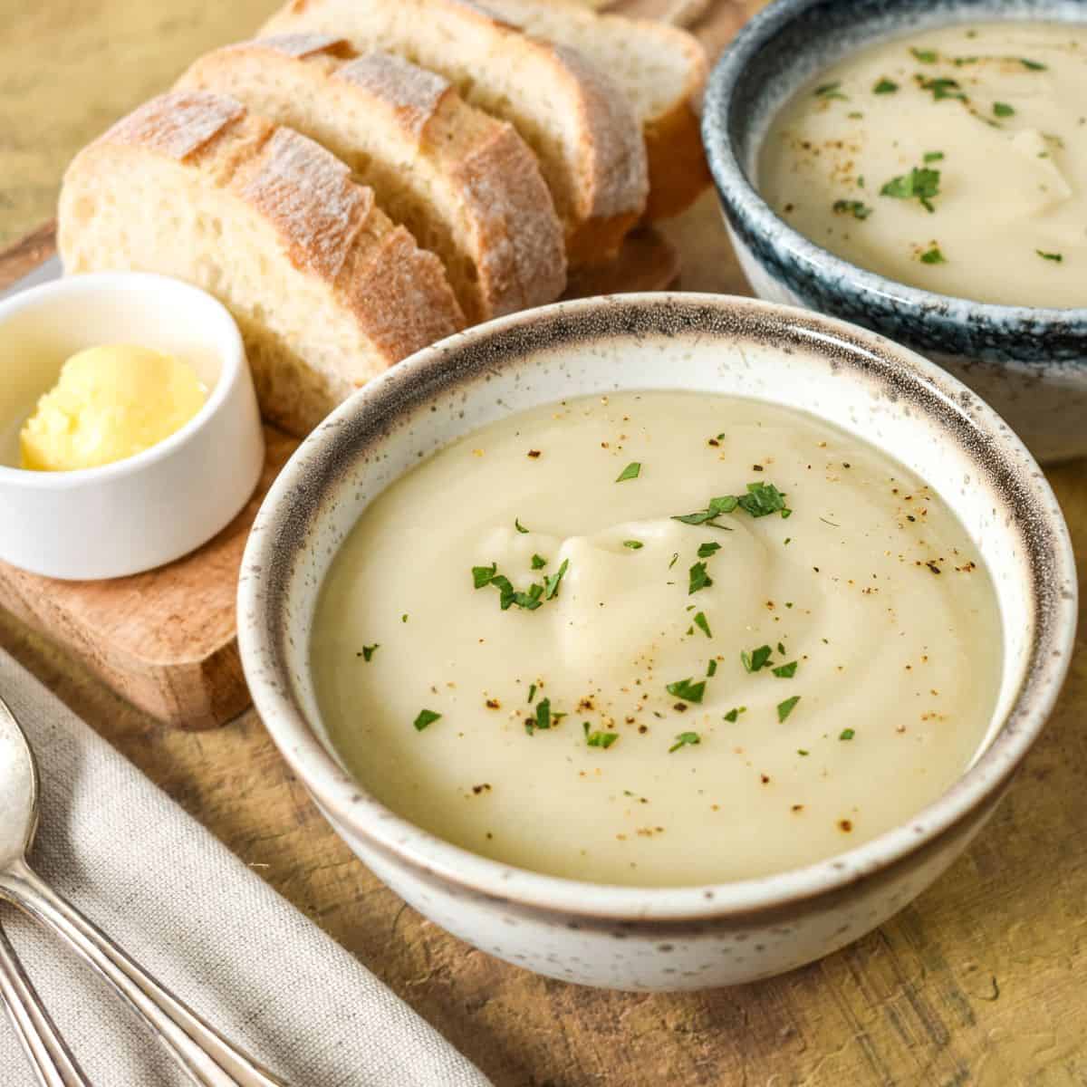 Close-up of a bowl of creamy soup topped with chopped parsley, with bread and butter in the background.