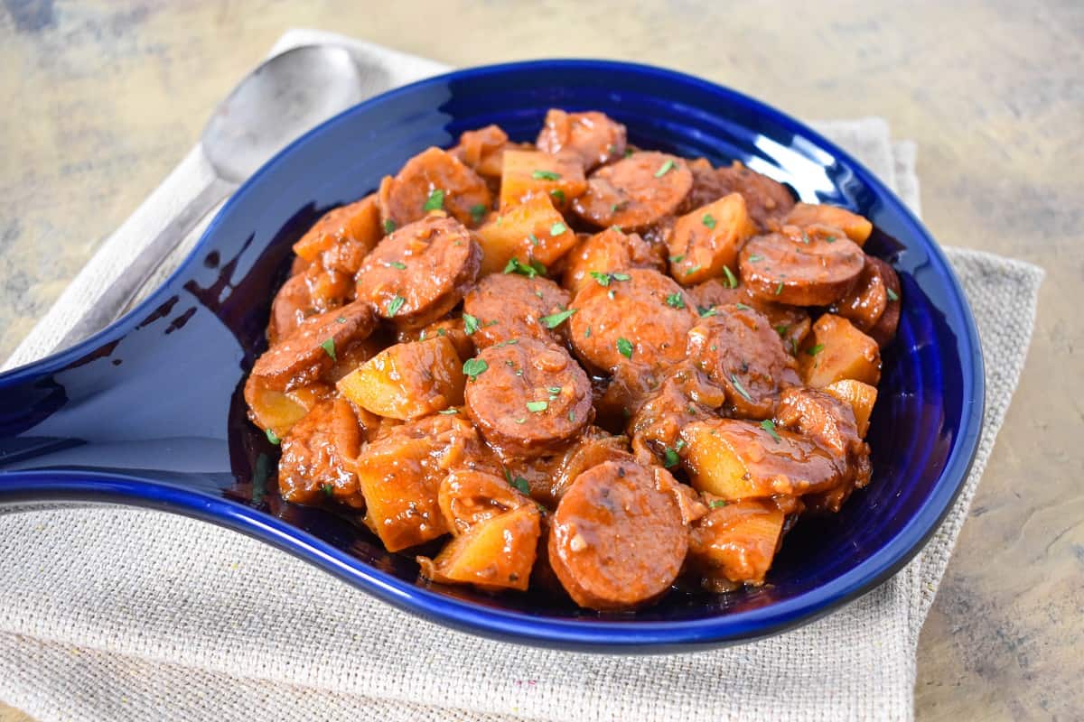 The smoked sausage and potatoes served in a blue crock and set on a beige linen.