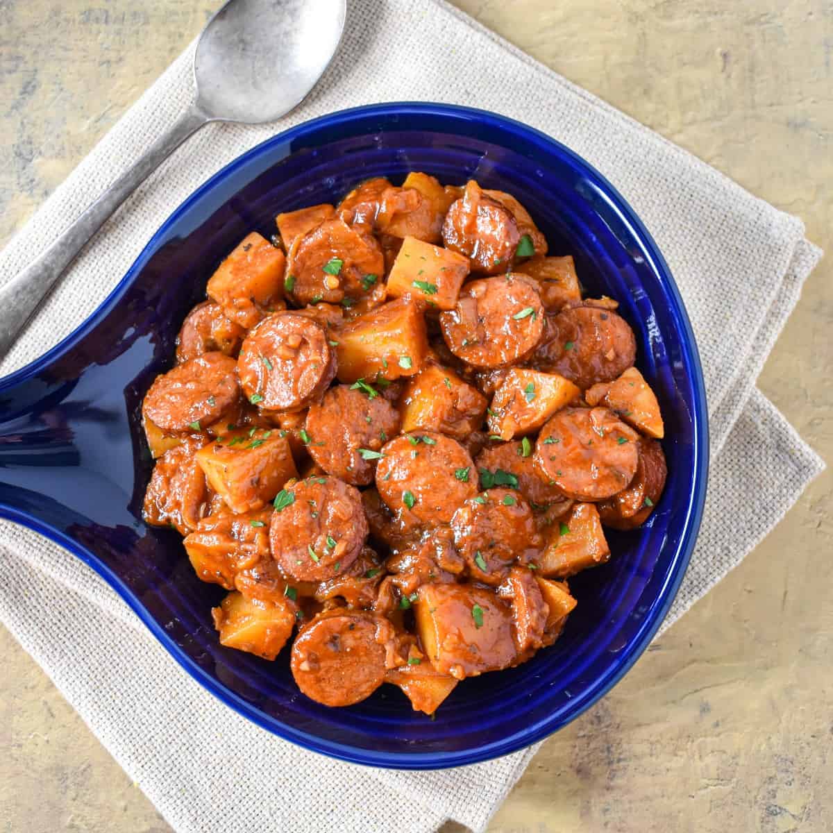 The smoked sausage and potatoes served in a blue crock and set on a beige linen.