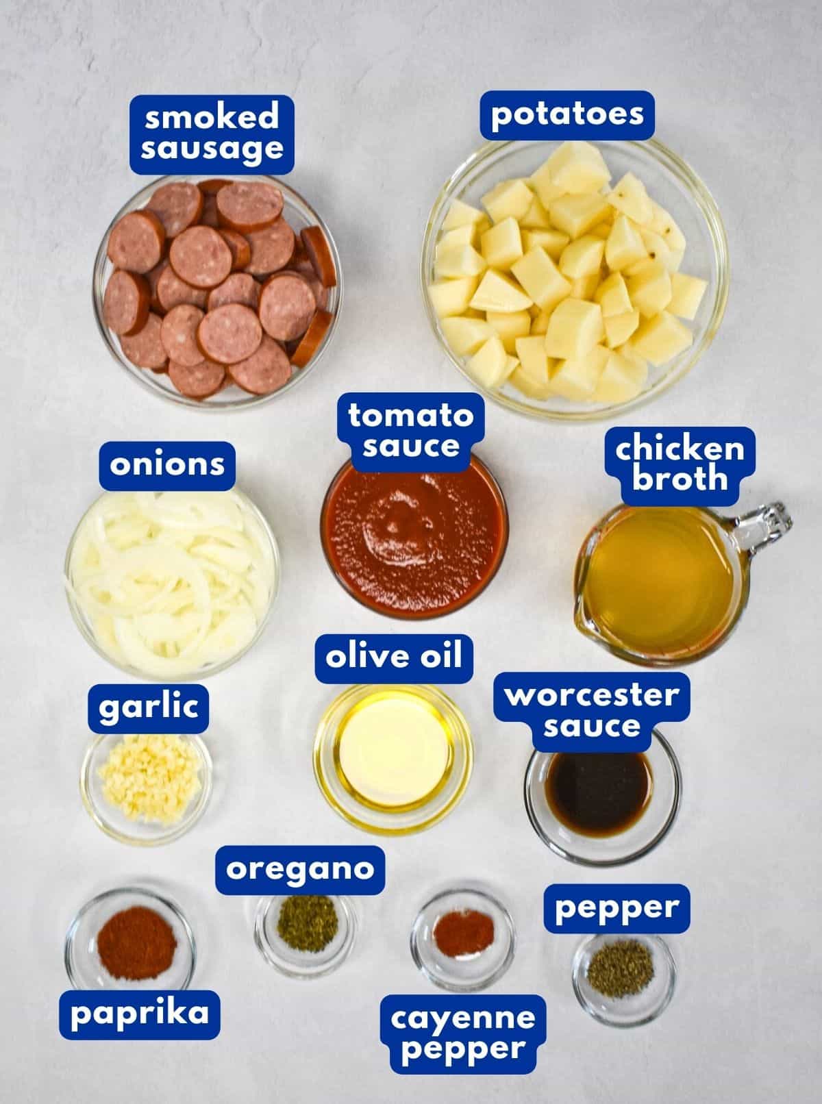 The ingredients for the sausage and potatoes skillet prepped and arranged on a white table with each labeled with blue and white letters.