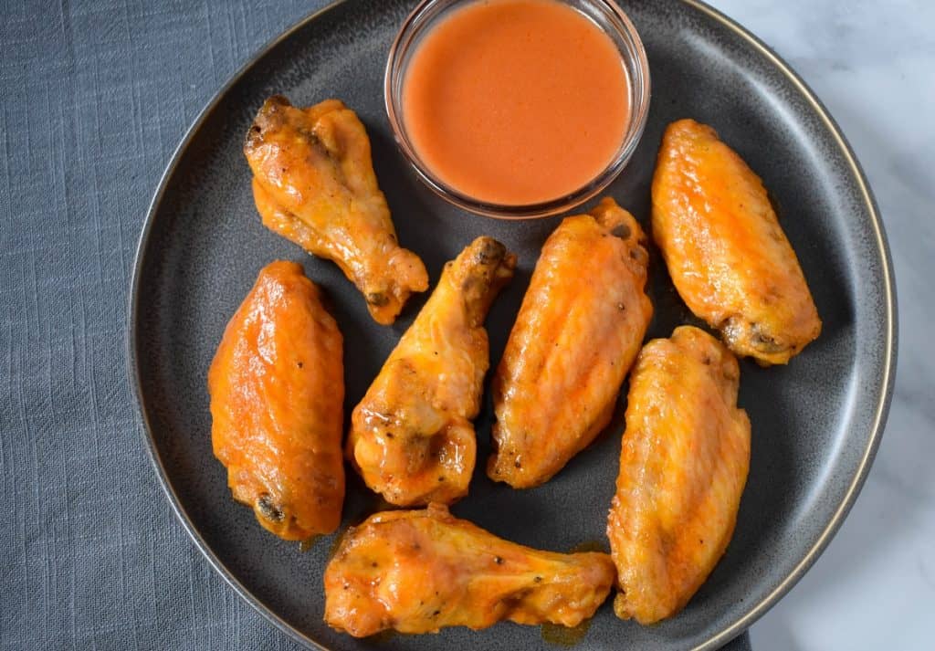 Seven chicken wings arranged on a gray plate with a side of buffalo sauce in a small bowl.
