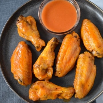 Seven chicken wings arranged on a gray plate with a side of buffalo sauce in a small bowl.
