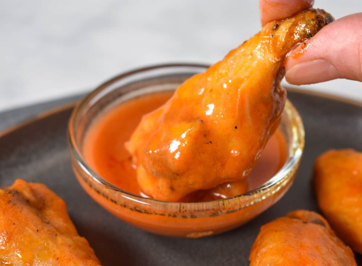A close up image of a drumette being dipped in a small bowl of buffalo sauce.