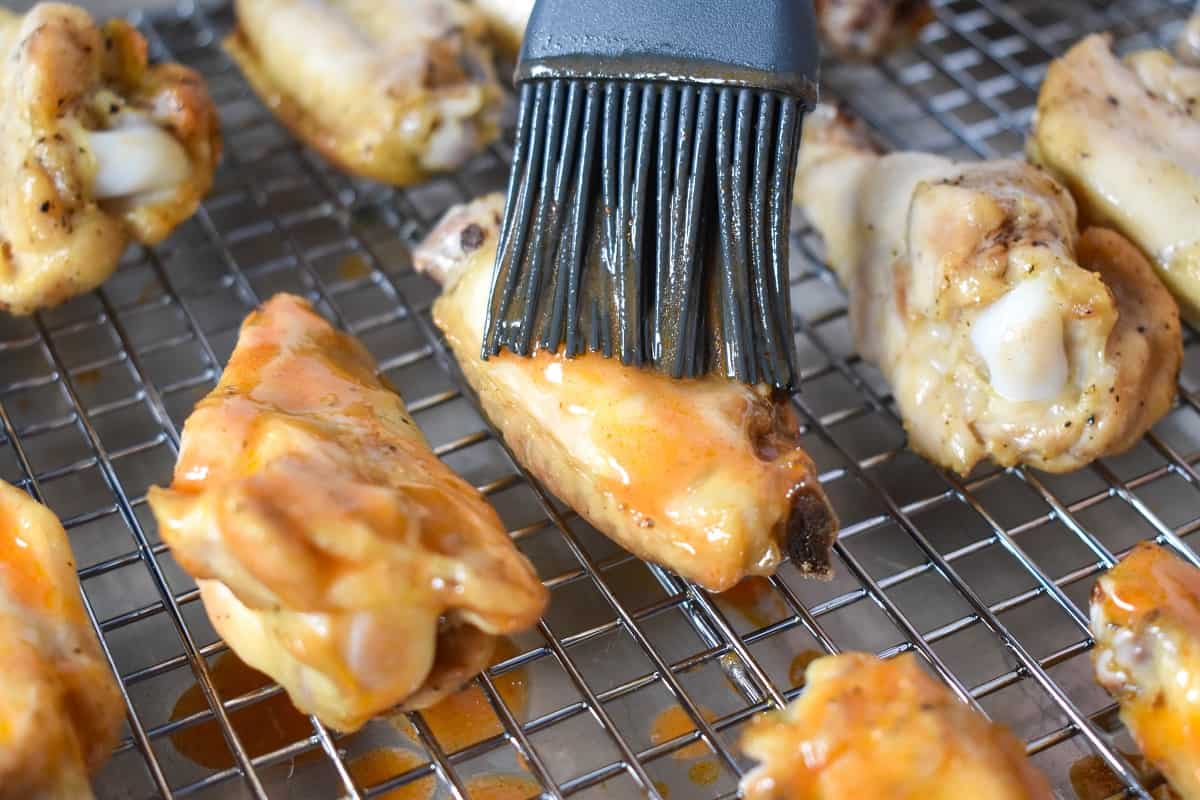 A close up of a few wings, one of which is having buffalo sauce applied with a gray pastry brush.
