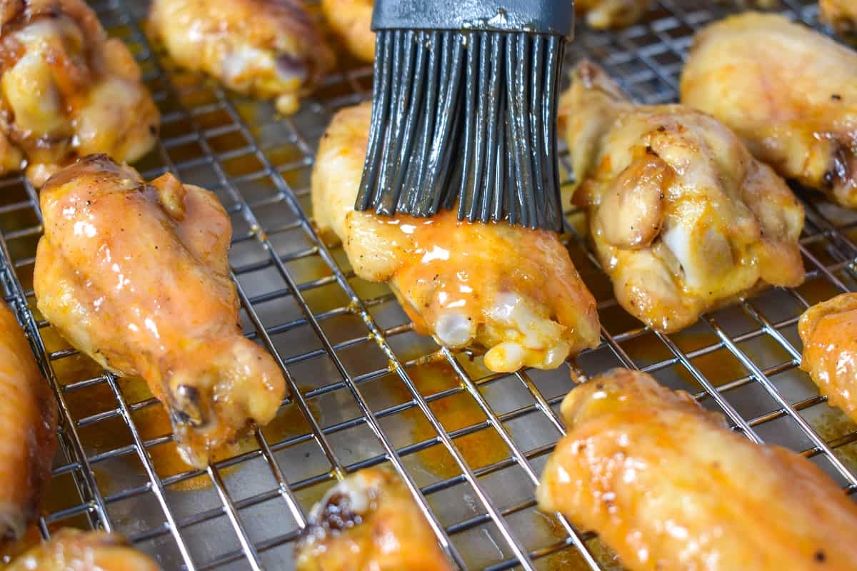 A close up of a few wings, one of which is having buffalo sauce applied with a gray pastry brush.