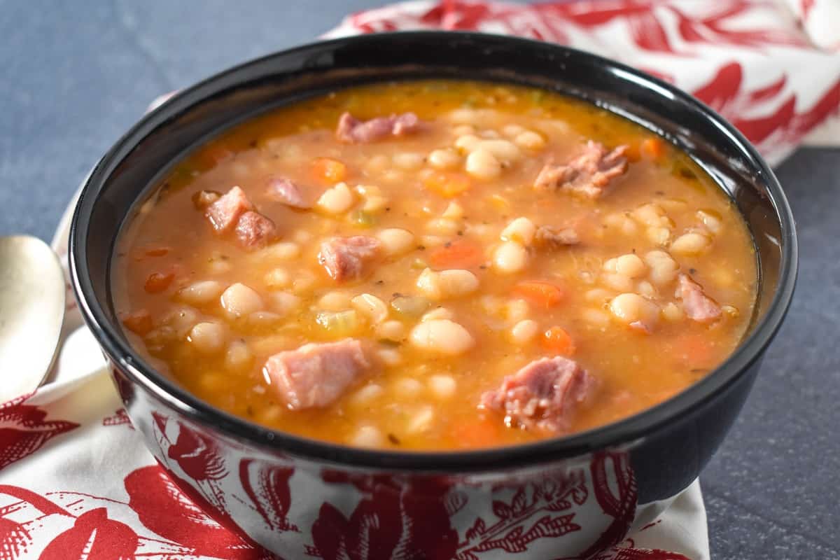 Navy bean soup served in a black bowl, with a red and white cloth napkin and spoon on the side.