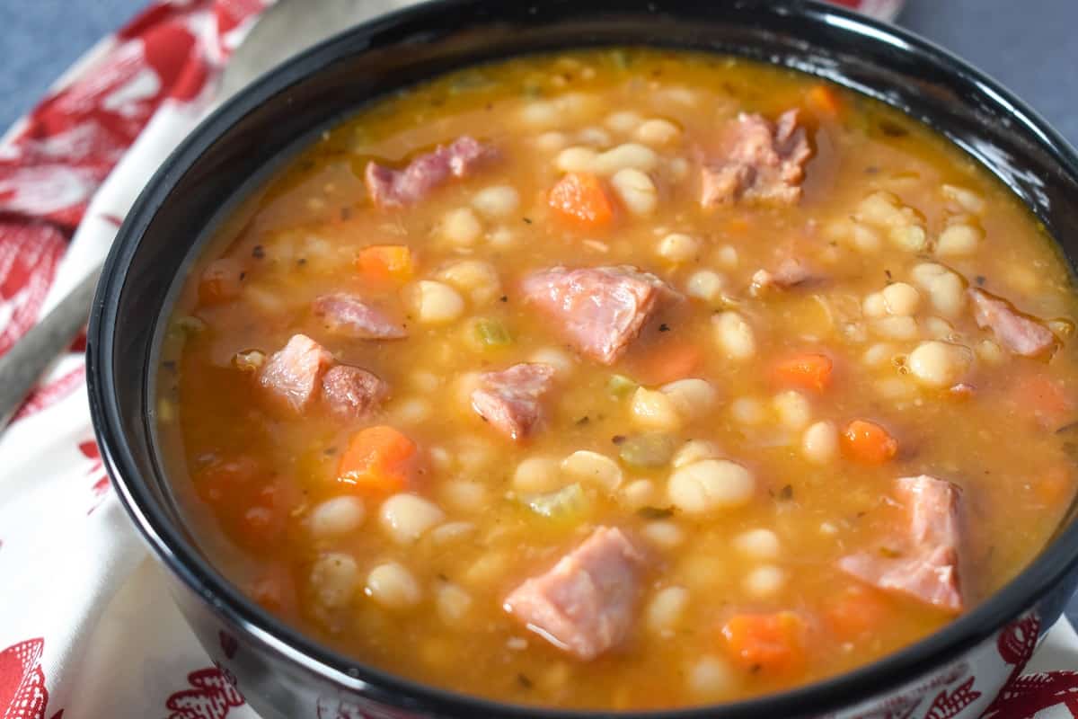 A close up image of navy bean soup served in a black bowl, with a red and white cloth napkin and spoon on the side.