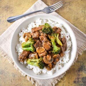 Bowl of beef and broccoli with mushrooms served over white rice, placed on a beige cloth with a fork in the background.