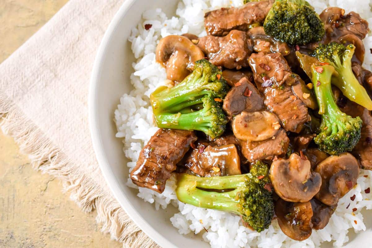 Close-up of beef, broccoli, and mushrooms coated in sauce over white rice in a shallow bowl.
