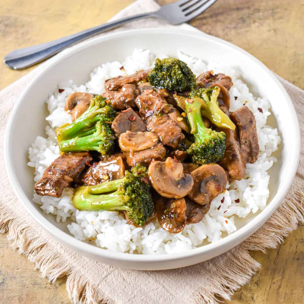Angled view of beef, broccoli, and mushrooms served over rice in a white bowl on a fringed napkin.