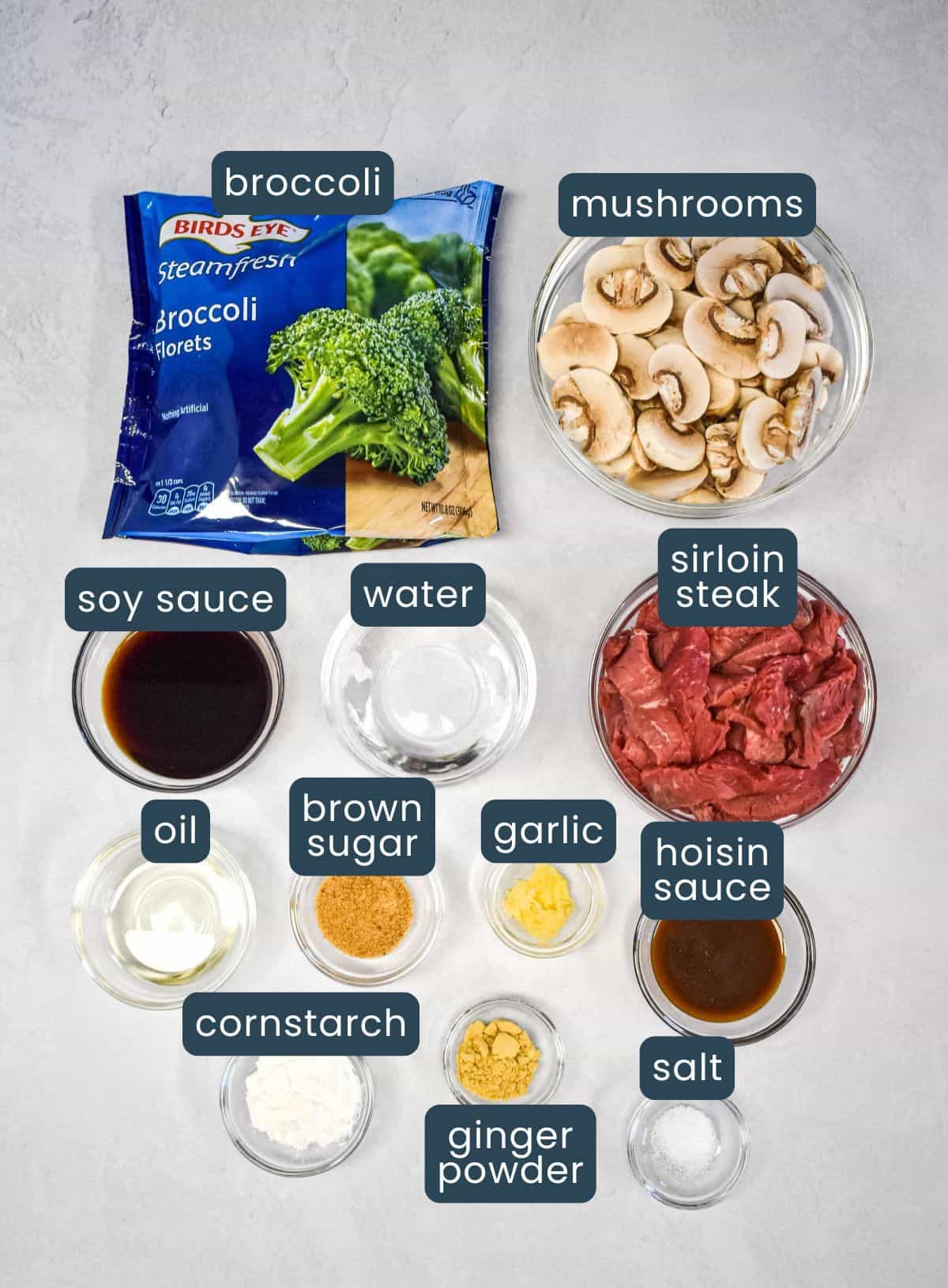 Overhead view of labeled ingredients in small bowls and containers on a light surface, including sliced mushrooms, frozen broccoli, sirloin steak, soy sauce, water, oil, brown sugar, garlic, hoisin sauce, cornstarch, ginger powder, and salt.