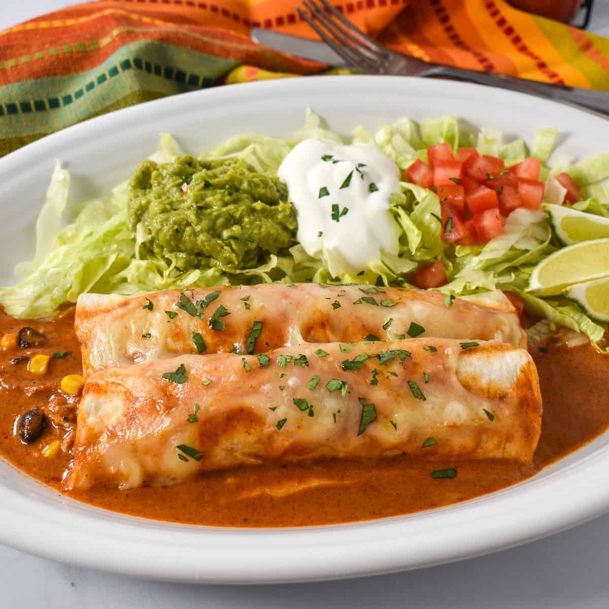 An image of the chicken enchiladas served on a large white plate with lettuce, guacamole, sour cream and tomatoes. In the background there is a linen with red, orange and green lines.