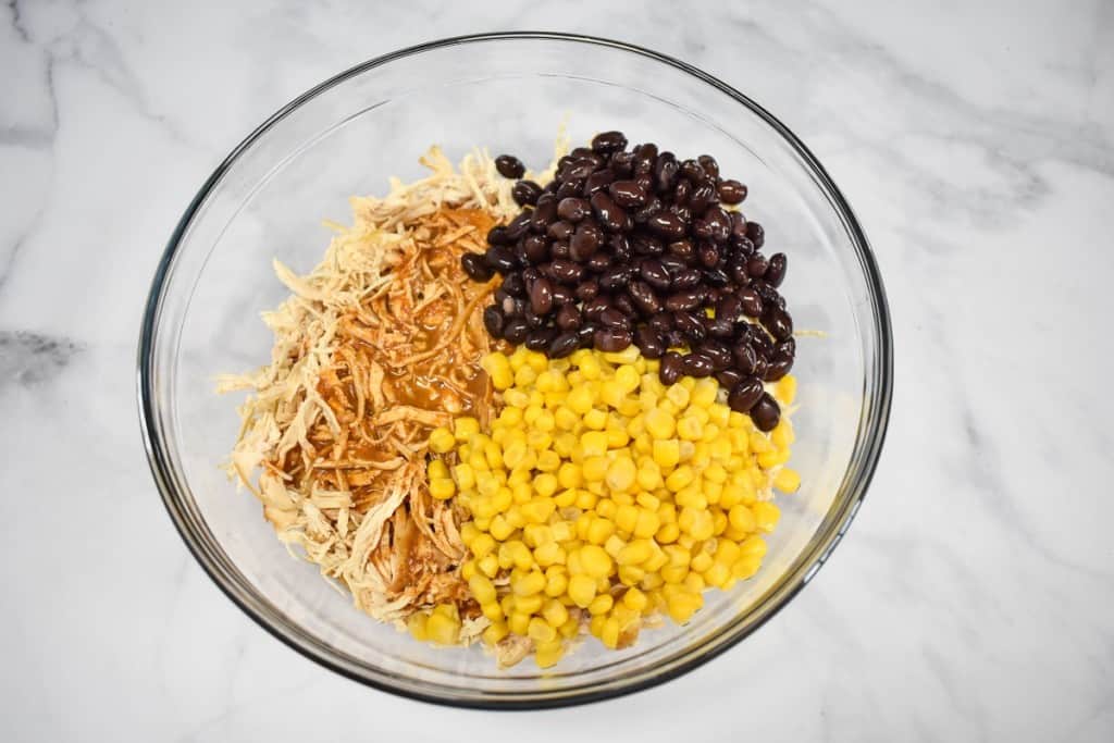 An image of shredded chicken, black beans and corn in a large glass bowl set on a white table.