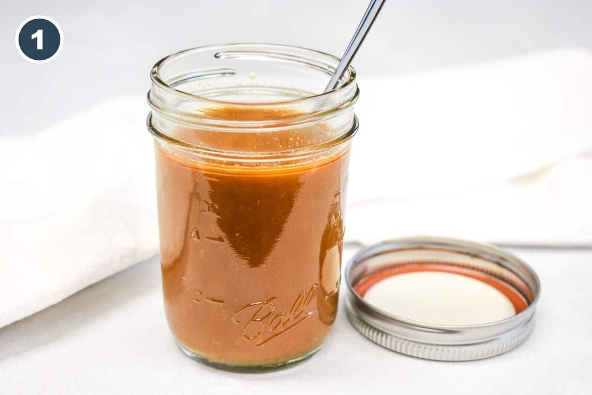 A jar filled with brown stir fry sauce and a spoon inside, with the lid off and resting beside it.