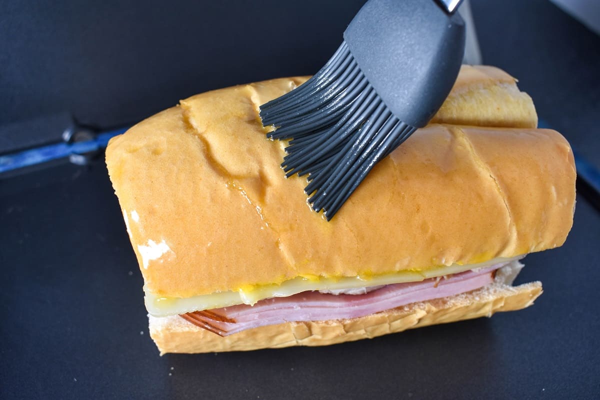 A gray silicone brush applying butter on the top slice of cuban bread.