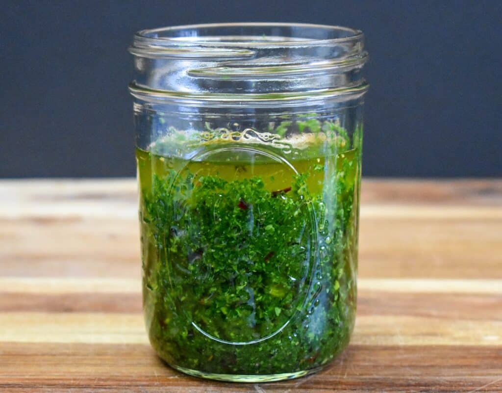 Chimichurri sauce in a clear glass canning jar displayed on a wood cutting board.