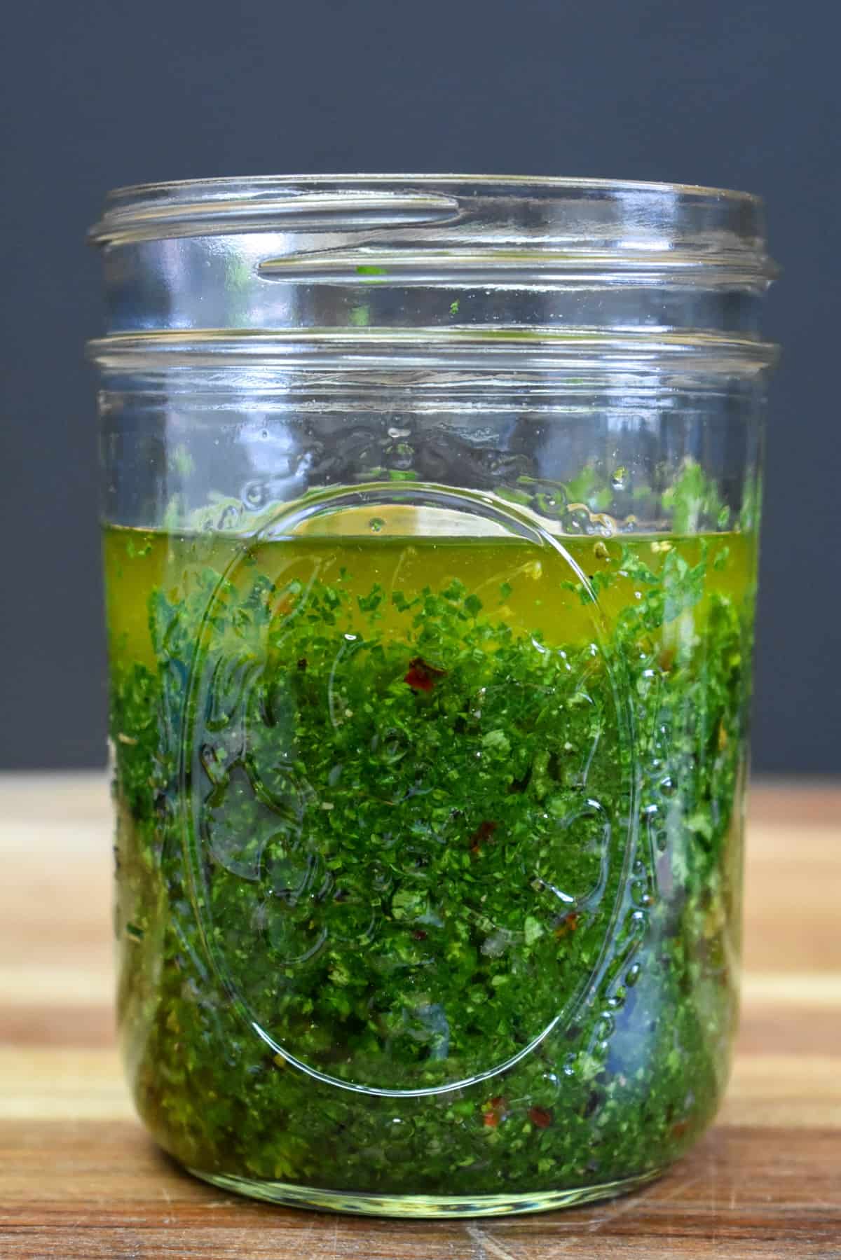 Chimichurri sauce in a clear canning jar displayed on a wood cutting board.