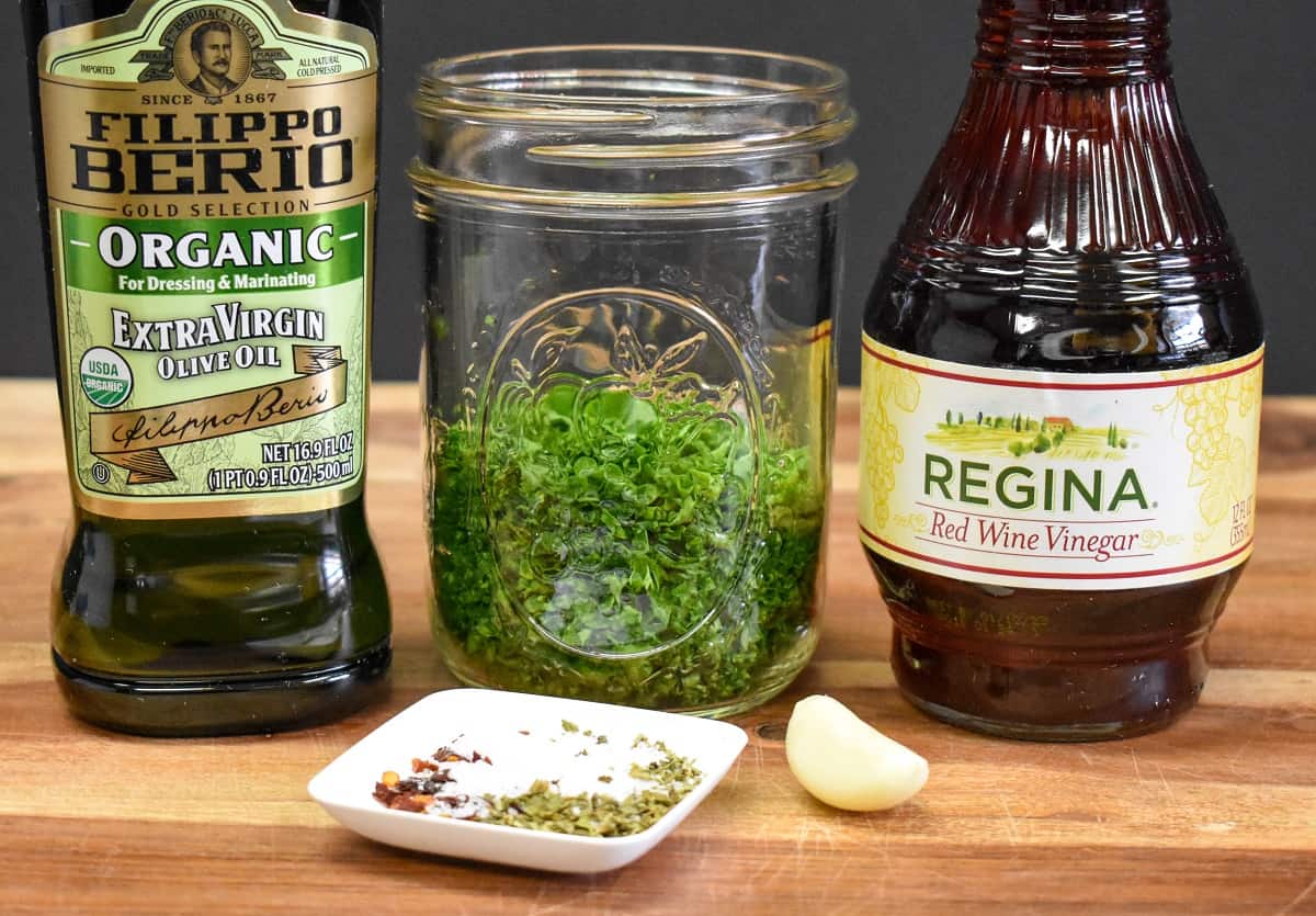 A bottle of olive oil, chopped parsley in a glass canning jar, a bottle of red wine vinegar, spices and a garlic clove arranged on a wood cutting board.
