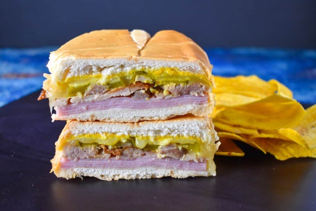 An image of the sandwich one half stacked on the other half with plantain chips on the right side and displayed on a black cutting board on a blue table.