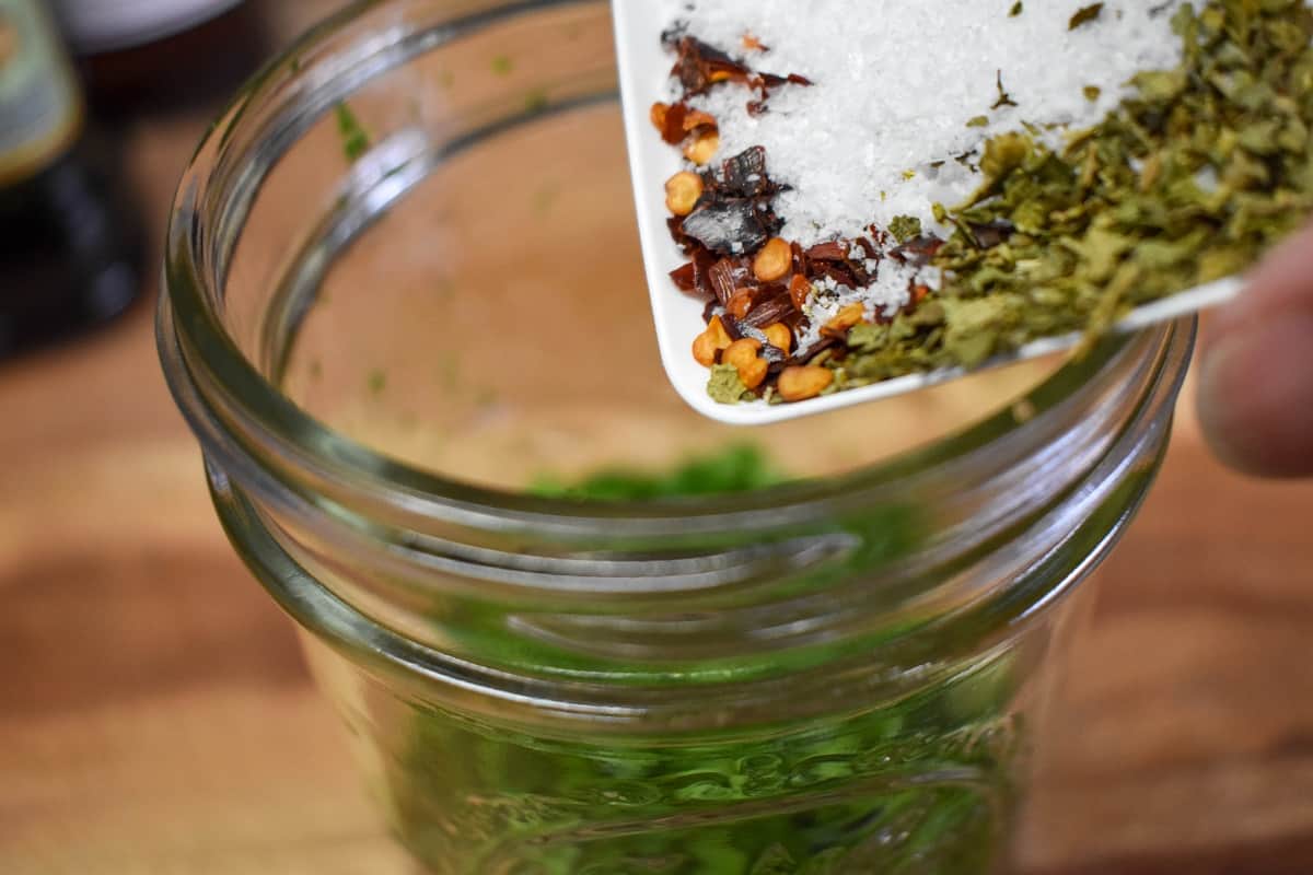 Salt, pepper, dried oregano, and red pepper flakes in a small white plate being added to the parsley in a glass canning jar.