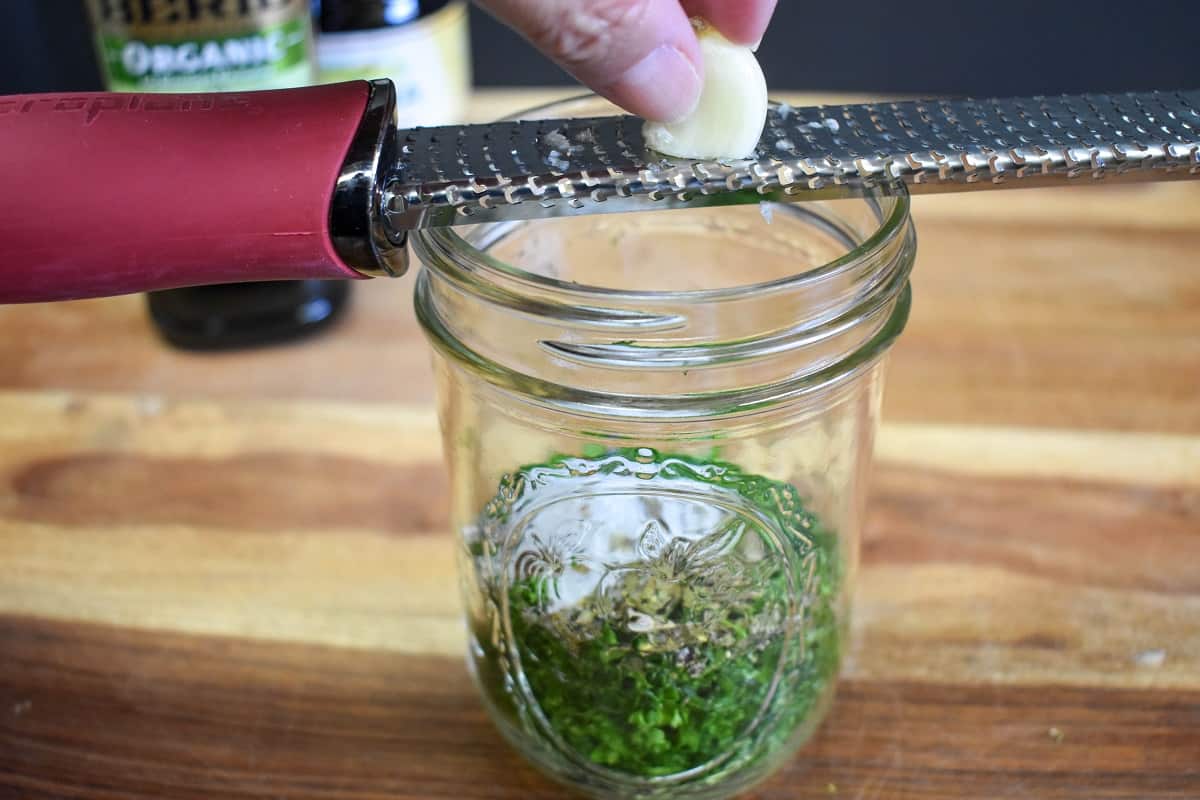 A garlic clove being grated using a hand held grater into a canning jar that contains chopped parsley and spices.