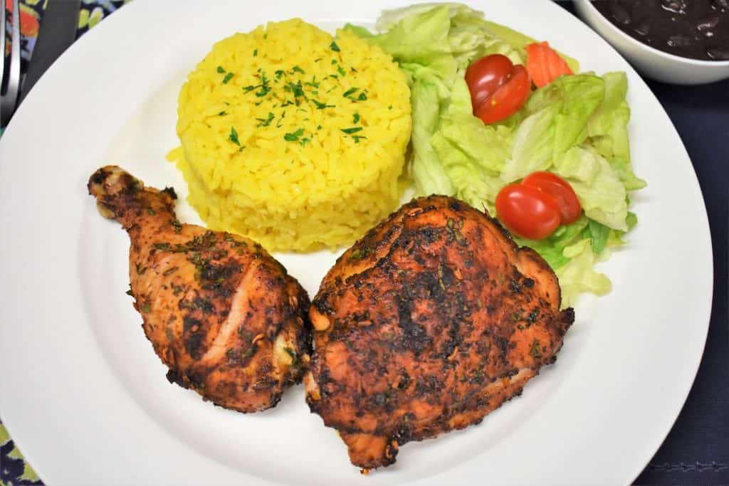 A cooked spicy baked chicken thigh and drum on a white plate with a side salad and yellow rice.