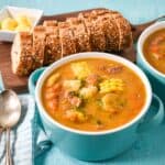 A bowl of Cuban ajiaco soup with corn, root vegetables, and meat, served with sliced bread and butter.