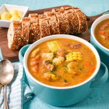 A bowl of Cuban ajiaco soup with corn, root vegetables, and meat, served with sliced bread and butter.