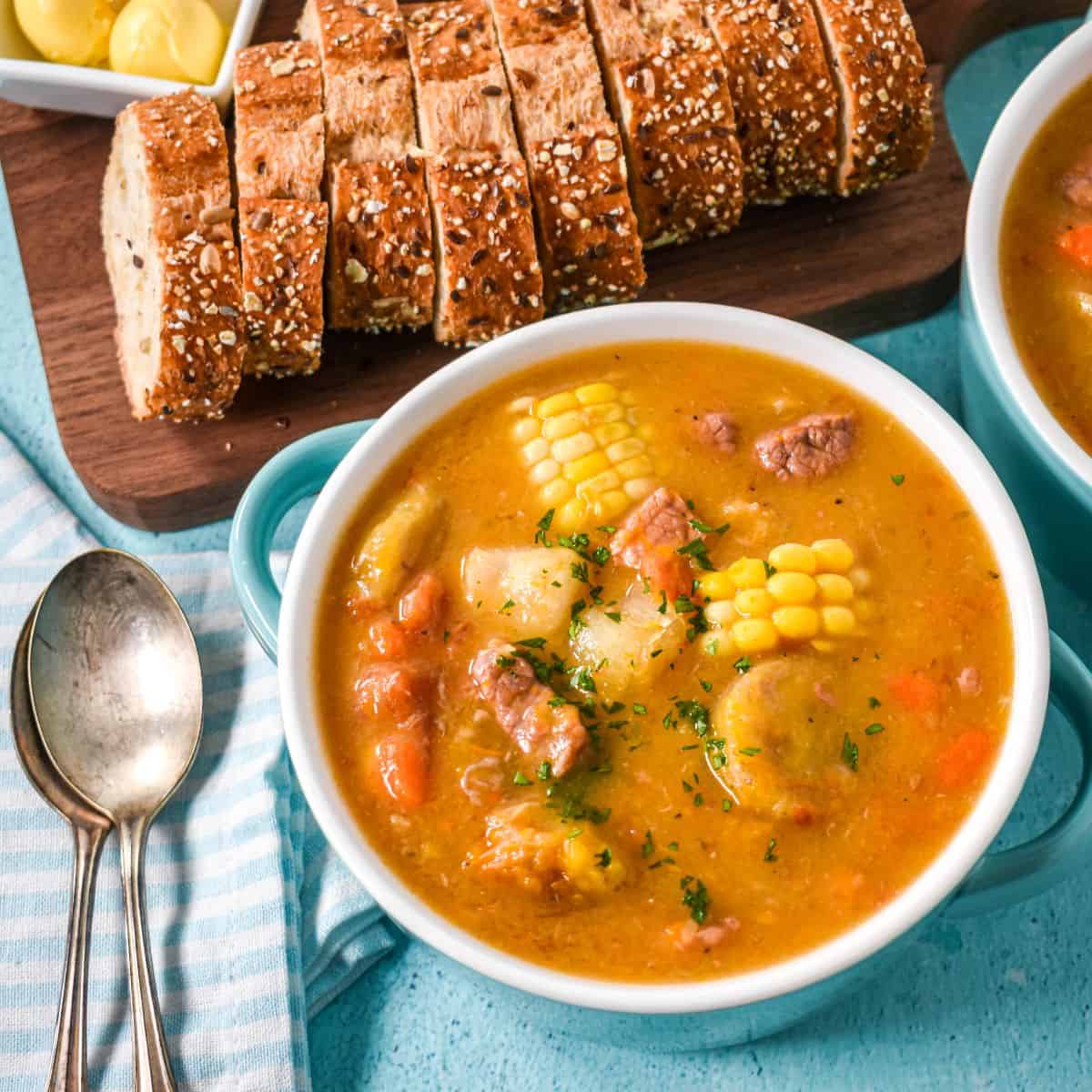 A bowl of hearty soup with corn, pork, yuca, and vegetables, served with whole grain bread.