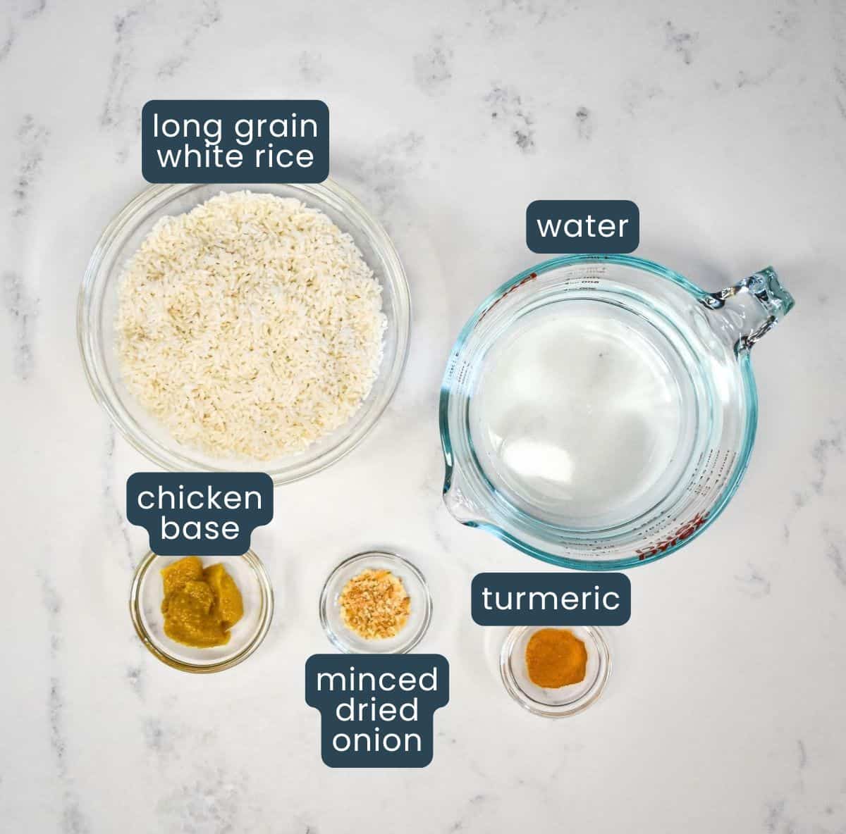 Ingredients for easy yellow rice: long-grain white rice, water, chicken base, minced dried onion, and turmeric set on a white table with each one labeled.