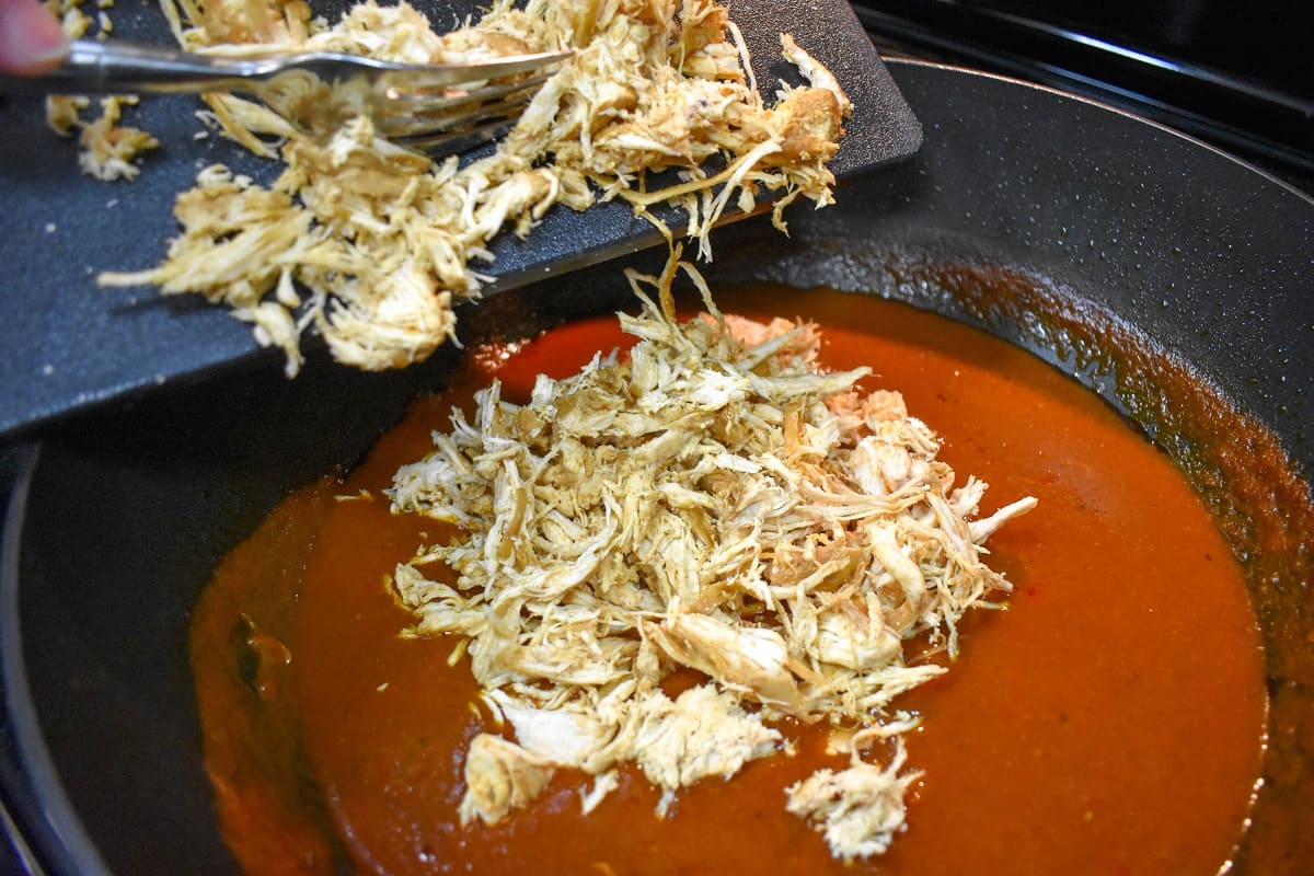 Shredded chicken being added to taco sauce in a large, black skillet.
