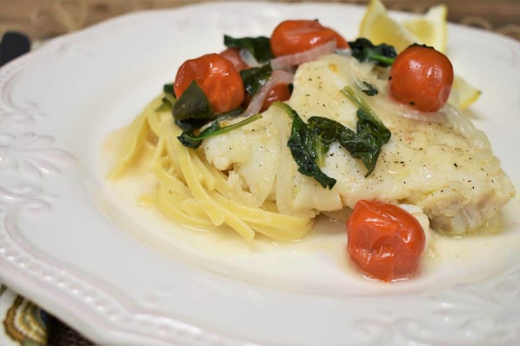 Cod, tomatoes and spinach served over pasta on a white plate.